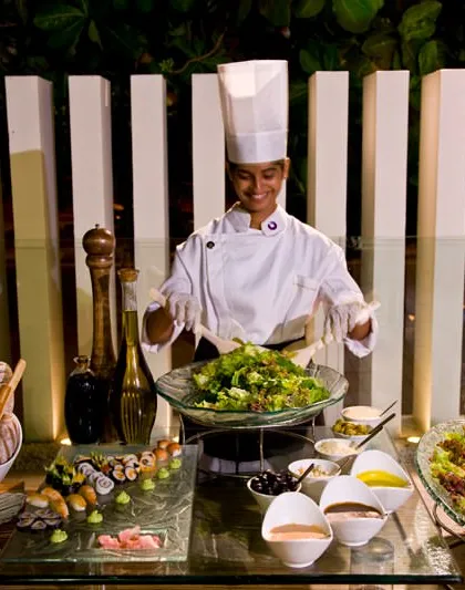 A smiling chef presents a freshly tossed salad at a vibrant salad bar. This setup includes a variety of fresh greens, toppings, dressings, and even vegetarian sushi, offering a healthy and customizable option for guests.