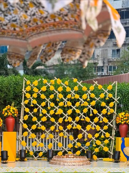 The Haldi stage setup, complete with a large printed umbrella providing shade. The combination of the traditional marigold backdrop with modern printed textiles creates a fresh and stylish look.