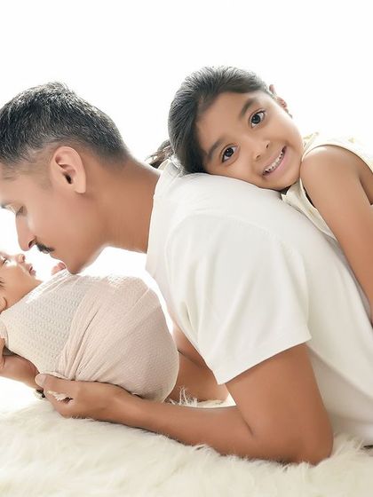 A creative and loving pose with the father, newborn, and older sister. The father kisses his new baby while the big sister leans on his back, creating a unique and memorable family portrait.