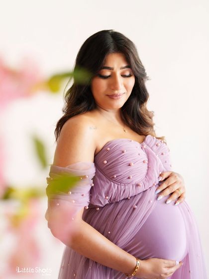 A soft and gentle close-up of a mother-to-be in a lavender gown. The focus is on her serene expression as she looks down, with a soft blur of flowers in the foreground.