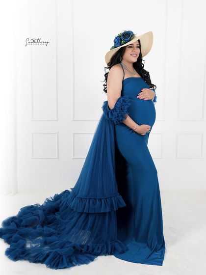 A stylish portrait of a mother-to-be in a deep blue gown, accessorized with a chic hat. The simple white background keeps the focus on her elegant and modern look.