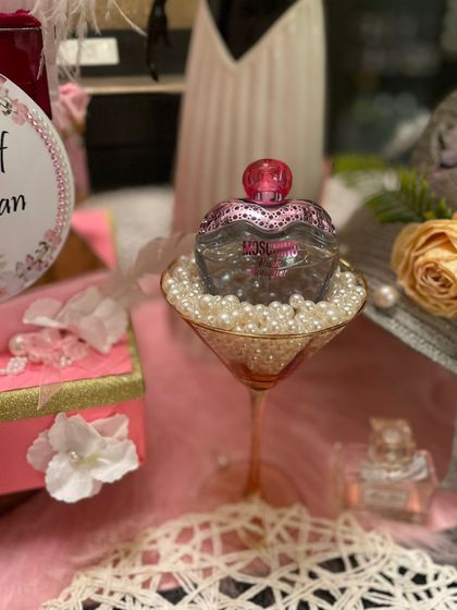 A detail from the perfume-themed table, where a bottle of Moschino perfume is displayed in a martini glass filled with pearls. It's a quirky and glamorous touch that adds personality to the decor.