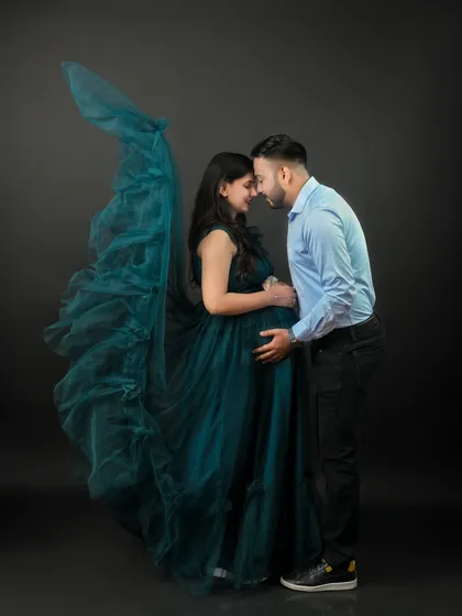 An intimate and elegant studio portrait for an expecting couple. The dark background and flowing teal gown create a sophisticated, modern look that focuses entirely on their connection.