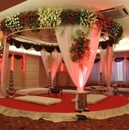 A circular mandap design for an indoor wedding. This setup uses a combination of red and white flowers on the canopy, with elegant white drapes and traditional low seating (baithak) for the ceremony.