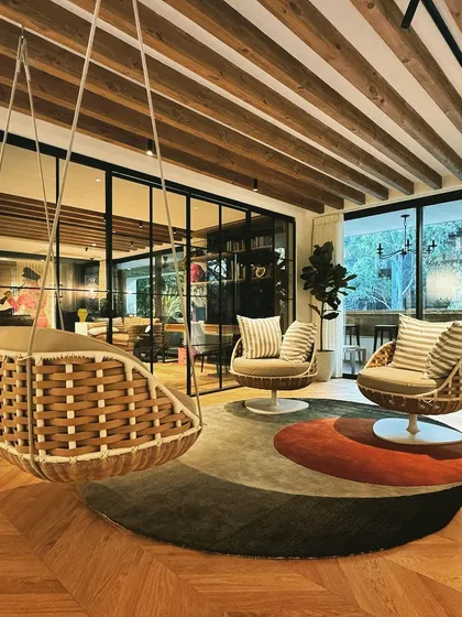 We created this informal lounge area as a peaceful escape from the formal settings of the house. A woven hanging swing chair and comfortable swivel chairs are arranged on a circular rug, encouraging relaxation and conversation. The space is defined by its connection to the outdoors and its biophilic design elements, making it a perfect spot to unwind.