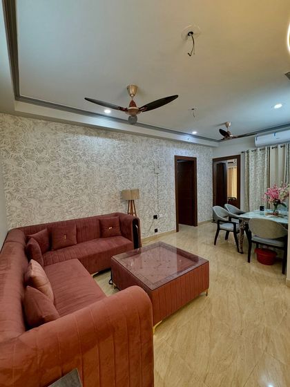 A full view of a living and dining area. The space features a textured wallpaper, a simple false ceiling, and a comfortable L-shaped sofa.