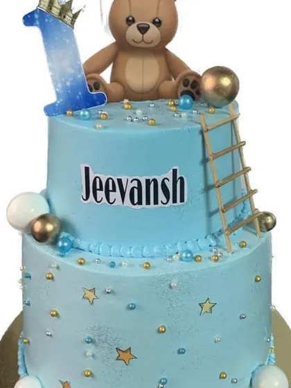 A cute blue two-tier cake with a teddy bear and ladder for a first birthday.