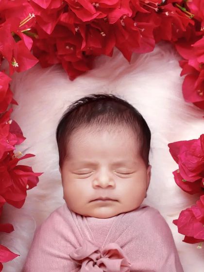 A close-up portrait of a baby framed by vibrant red bougainvillea flowers, creating a beautiful contrast with their peaceful expression.