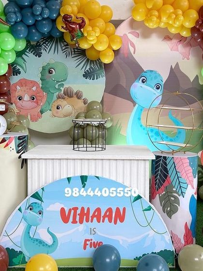 A cute and colorful dinosaur-themed 5th birthday party. This setup includes a backdrop with friendly dinosaurs, character cutouts, and a vibrant balloon garland.