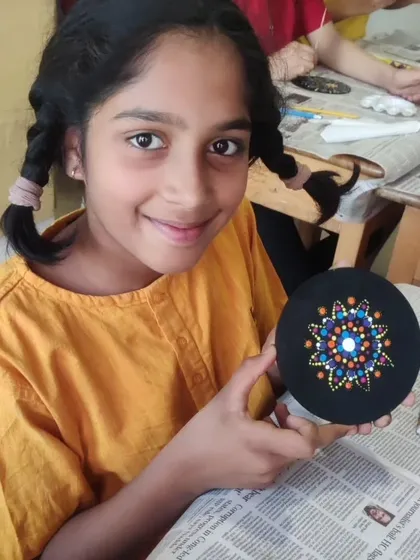 A young artist proudly holds up her dot art coaster, which features a star-like pattern in shades of blue and orange. Each piece from the workshop is a small masterpiece.