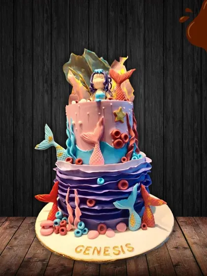 This two-tier mermaid cake is an underwater fantasy. The purple and blue design features multiple mermaid tails, coral, and a beautiful mermaid figure on top, creating a magical ocean scene.