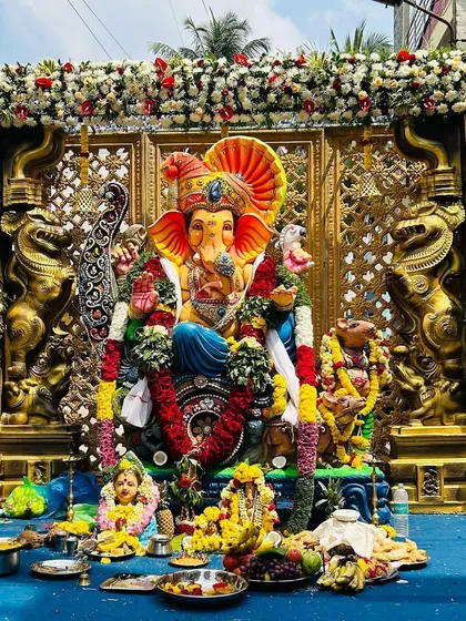 A grand Ganesh Chaturthi pandal we created for a community celebration. The setup features a majestic Ganesha idol set against an ornate gold lattice backdrop, flanked by traditional Yali pillars and adorned with lush garlands of white and red flowers.