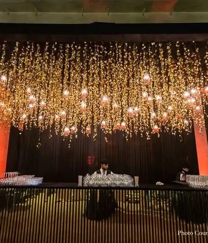 This modern bar counter features a black slatted wood design. The space above is decorated with a stunning installation of hanging flowers and lights, creating a dramatic and sophisticated look for an evening event.