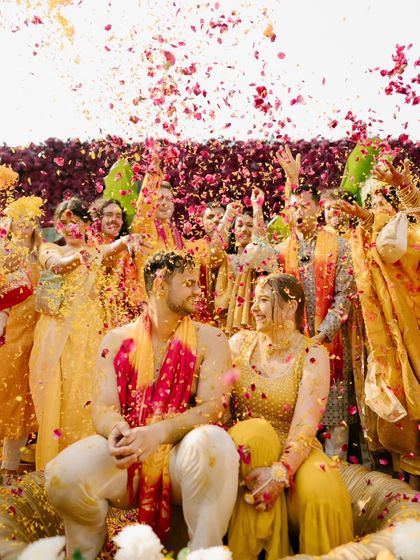 The couple sits together, surrounded by family and a storm of pink and yellow petals, a perfect summary of a joy-filled Haldi.