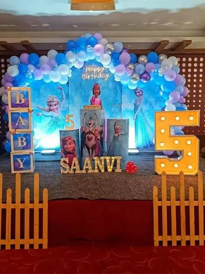 A complete stage setup for a Frozen-themed 5th birthday, featuring character standees, a balloon arch, and light-up numbers.