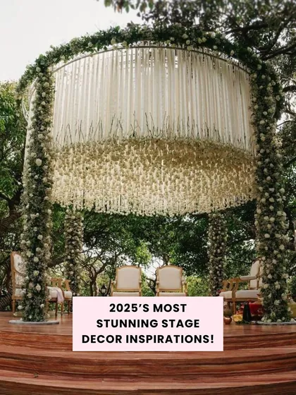 A stunning circular mandap design with a canopy of hanging white ribbons and flowers, set on a wooden stage amidst lush greenery for a truly unique outdoor ceremony.