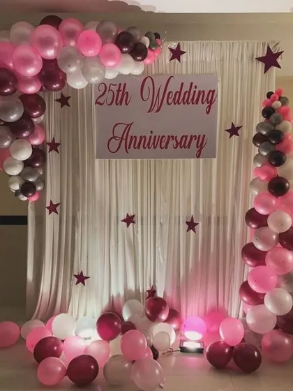 A classic 25th wedding anniversary backdrop with a white curtain and a sign. The setup is framed by a balloon arch in shades of pink, maroon, and white, with star cutouts adding a whimsical touch.