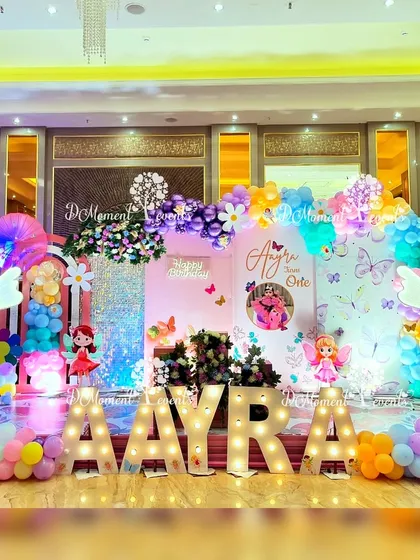 A vibrant fairy and butterfly themed first birthday stage for Aayra. The decor features marquee letters, a sequin backdrop, colorful balloon garlands, and cute fairy cutouts, all coming together to create a cheerful and magical setting.