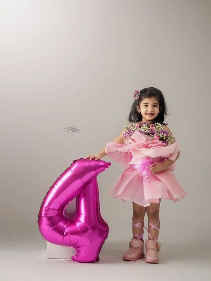 Celebrating turning four with a pretty pink theme. This little girl is dressed like a ballerina, holding a bouquet, next to a large number '4' balloon.
