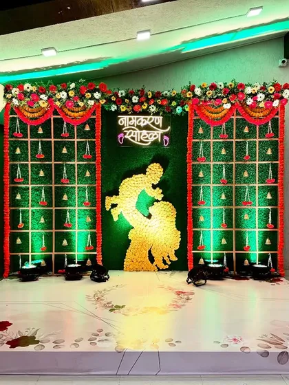 Honoring tradition with this beautiful Namkaran Sohala decor. The stage features a classic green backdrop with intricate genda phool (marigold) hangings and a central silhouette, creating a perfect setting for a traditional naming ceremony.