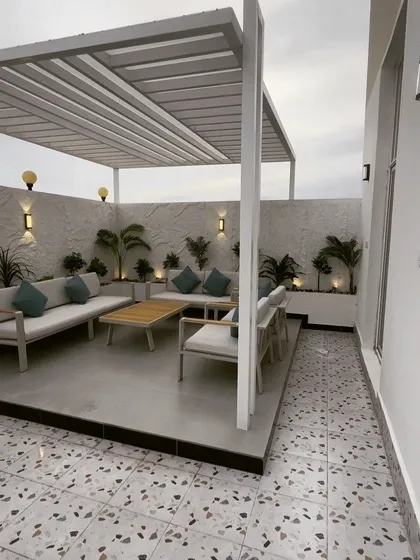 A bright and airy terrace design featuring a white metal pergola over a raised concrete platform. The space is furnished with modern outdoor sofas and planters, set against a textured white wall with accent lighting.