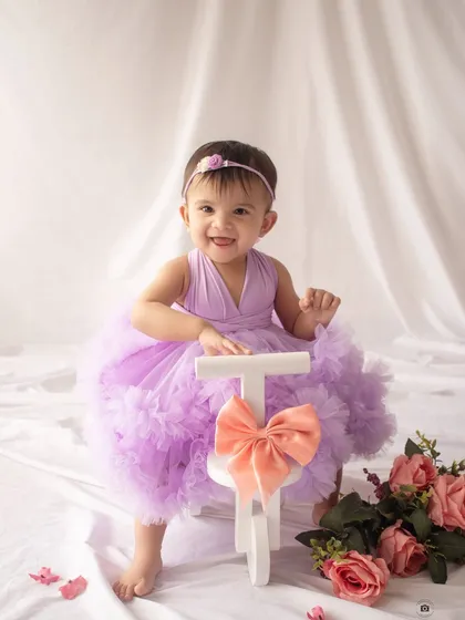 A joyful one-year-old in a lavender tutu, posing with a small white tricycle and a big smile.