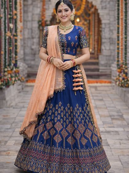 A beautiful royal blue silk lehenga paired with a contrasting peach dupatta. The skirt features traditional woven patterns, and the blouse has intricate multi-color embroidery on the sleeves.