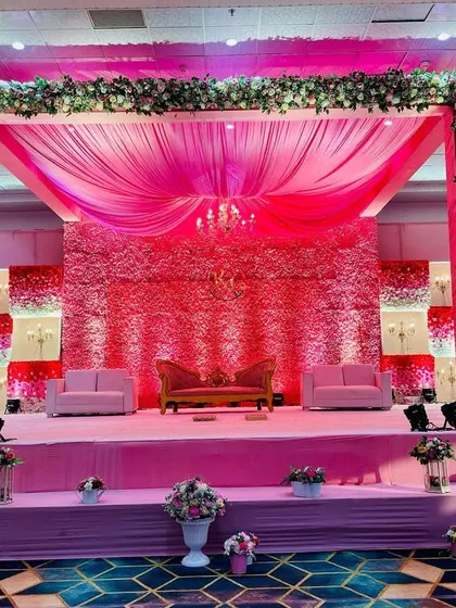 A massive, grand stage we designed for a wedding reception with a bold pink theme. The backdrop features a textured wall of pink flowers, complemented by draped fabrics, elegant sofas, and dramatic lighting to create a luxurious and romantic setting.
