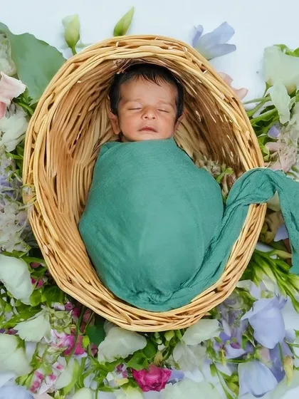 A newborn wrapped in green, sleeping in a basket surrounded by flowers. The overhead angle provides a beautiful view of the entire setup.