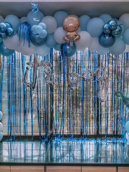 An 'It's a Boy' welcome decoration against a window, using a blue foil curtain as a backdrop for the balloon arch and props.