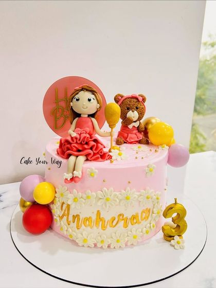 A pretty pink cake for a 3rd birthday, featuring a custom doll and teddy bear fondant topper. The daisy details add a sweet, delicate touch.