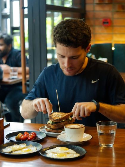 A customer enjoying a hearty breakfast with our stacked club sandwich, coffee, and sides of fried eggs and roasted tomatoes.