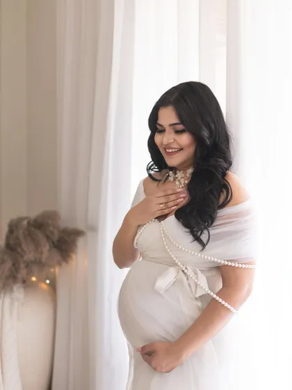 A candid, smiling portrait of the mother-to-be. The soft, natural light from the window and the delicate pearl accessories add to the angelic and serene feel of this shot.