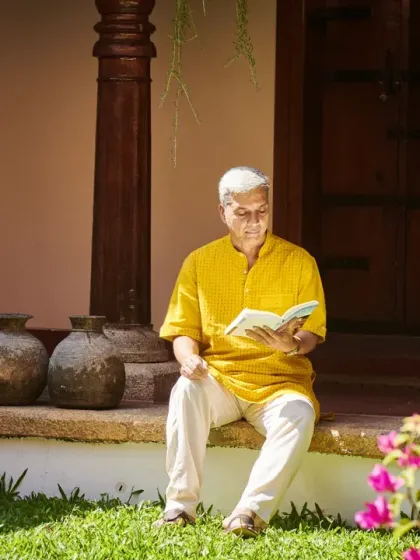A guest enjoys a book while sitting on the veranda of a traditional Nalukettu. Surrounded by the timeless charm of wooden pillars and a Tulsi plant, he embodies the calm and reflection our environment inspires.