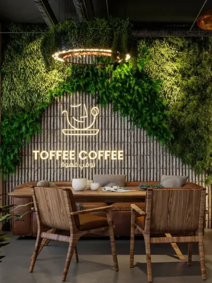 This cafe design features a prominent green wall as a backdrop for the brand's logo. The use of natural wood chairs and a leather sofa creates a comfortable, rustic, and earthy "Toffee Coffee" cafe vibe.
