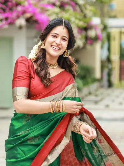 Bright and cheerful outdoor portraits of an actress in a vibrant green and red saree. The natural lighting and blooming flowers in the background complement her radiant smile.