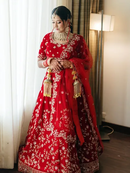 A full-length bridal portrait by the window. The soft, diffused light creates a classic and elegant look, highlighting the rich red of her lehenga.