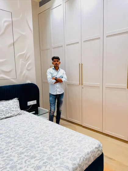 Standing in a newly completed bedroom. This space features a full-height white wardrobe with long brass handles and a bed with a deep blue upholstered headboard.