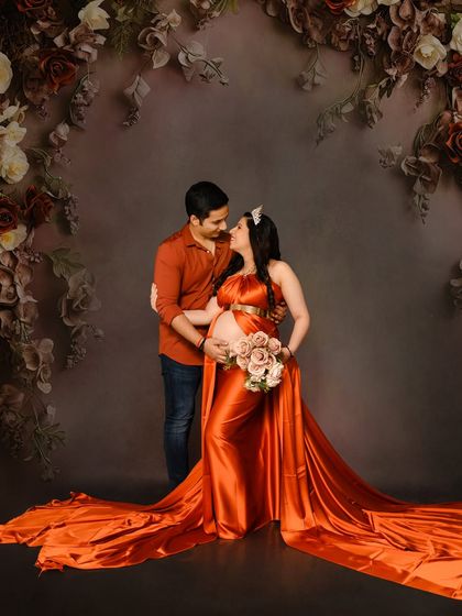 An intimate couple's portrait against a dark floral backdrop. The rich orange satin gown adds a vibrant pop of color and elegance.