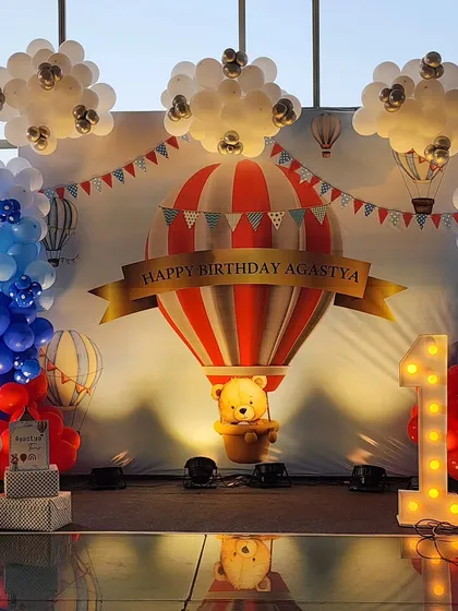 A grand first birthday celebration with a hot air balloon theme. This custom backdrop features a large balloon with a teddy bear, surrounded by balloon clouds and a lit-up "ONE" marquee sign, creating a perfect photo opportunity.