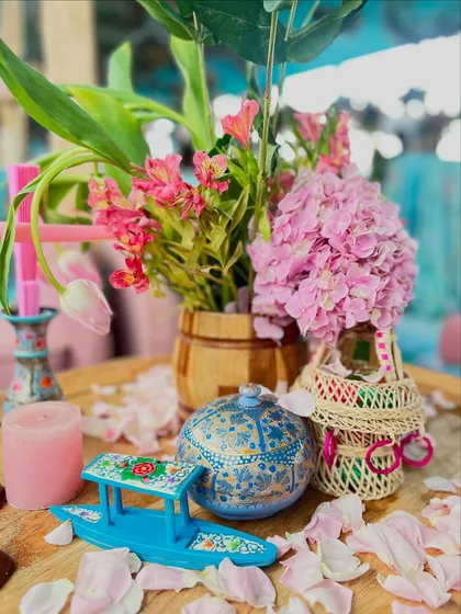 A close-up of the Kashmiri decor details, including a miniature, hand-painted shikara boat, a traditional papier-mâché box, and scattered rose petals.