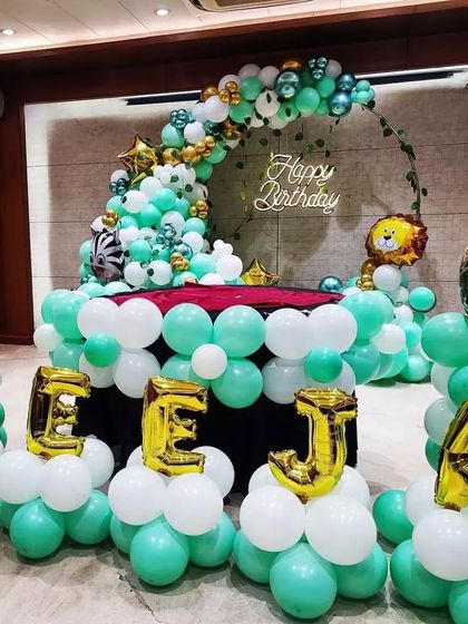 A jungle-themed party with personalized name balloons. The name "REEJA" is spelled out in gold foil balloons, set against a backdrop of green and white balloons with animal accents.