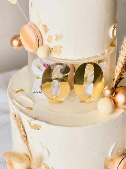 A sophisticated two-tier cake for a 60th birthday celebration in an ivory and gold theme. This design features a clear separator tier, macarons, and dried palm leaves for a modern, celebratory feel.