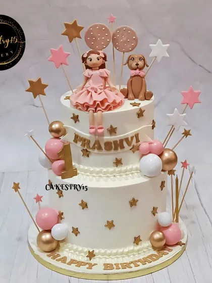 A charming two-tier doll-themed cake for a first birthday. This black forest cake is decorated in a pink and gold star theme, featuring a girl figurine and her pet dog.