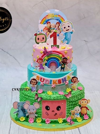 A magnificent three-tier Cocomelon cake for a first birthday. This design features a grass-like bottom tier, sparkling rhinestone ribbons, and all the main characters.