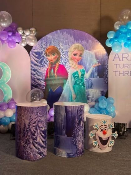 A 'Frozen' theme setup for a third birthday, with character backdrops, themed pedestals, and a balloon arrangement in shades of blue, purple, and white.