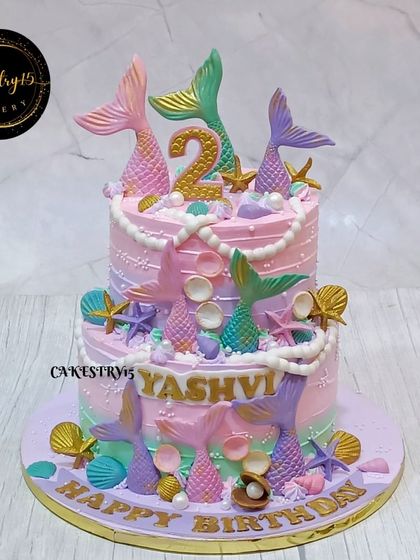 A whimsical two-tier mermaid-themed cake for a second birthday. This design features pastel pink and green frosting, with fondant mermaid tails, pearls, and seashells creating an underwater fantasy.