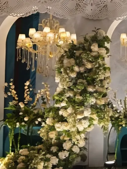 An elegant stage setup with cascading white floral arrangements flowing from teal-colored consoles, set against a white backdrop with chandeliers.