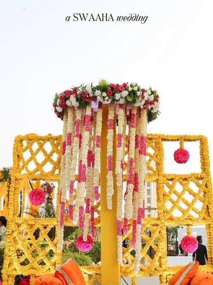 A beautiful floral installation for a Haldi ceremony, featuring a chandelier of jasmine and pink flowers, adding a touch of elegance to the vibrant yellow decor.