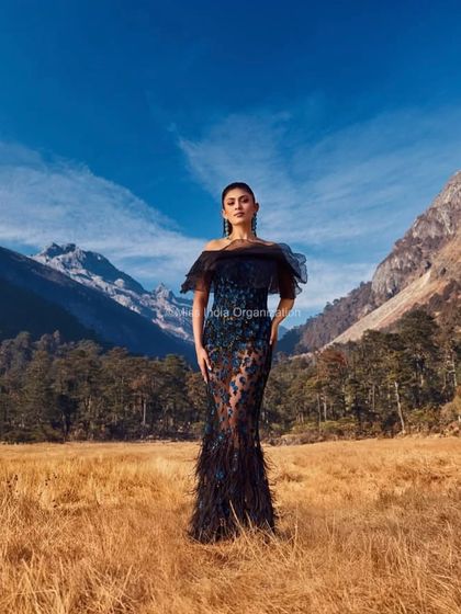 A dramatic editorial shot featuring a model in a stunning black gown against the majestic mountains of Arunachal Pradesh. Her hair is styled in a chic updo to accentuate her neckline and the dress's silhouette.